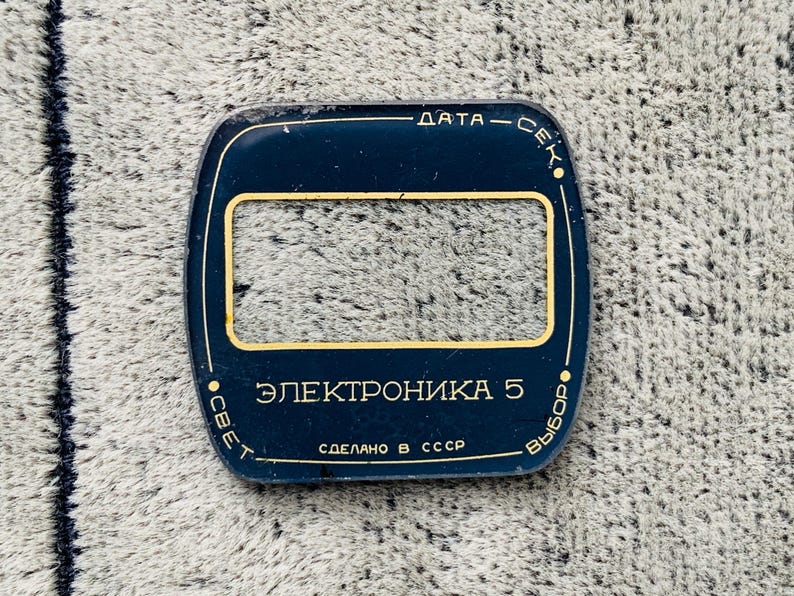 May include: A dark blue, rectangular watch face cover with a gold-colored rectangular window in the center. The text on the cover is in Cyrillic script, with the words "DATA - SEC", "ELECTROHIKA 5", and "MADE IN USSR".