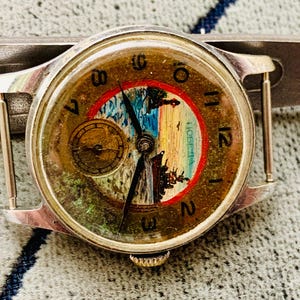 Puede incluir: Reloj de pulsera vintage con caja plateada y esfera redonda. La esfera del reloj presenta un dise&ntilde;o de paisaje con un borde rojo y una peque&ntilde;a subesfera. Las manecillas son negras y los n&uacute;meros est&aacute;n en una fuente cl&aacute;sica.