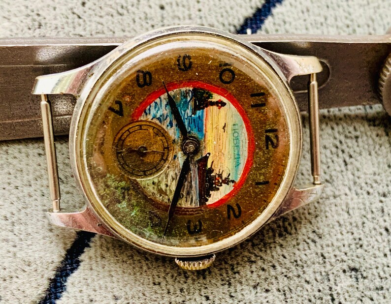 Puede incluir: Reloj de pulsera vintage con caja plateada y esfera redonda. La esfera del reloj presenta un dise&ntilde;o de paisaje con un borde rojo y una peque&ntilde;a subesfera. Las manecillas son negras y los n&uacute;meros est&aacute;n en una fuente cl&aacute;sica.