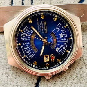 May include: A silver Orient automatic wristwatch with a dark blue face. The watch has gold hands, hour markers, and a date display. The face includes text indicating it is a 21-jewel, multi-year calendar watch.