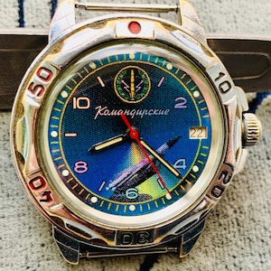 Op de afbeelding: Een vintage polshorloge met een zilverkleurige kast en een blauwe wijzerplaat. De wijzerplaat toont het Cyrillische woord "Командирские" en een raketafbeelding. Het horloge heeft een rode secondewijzer en lichtgevende uurmarkeringen.