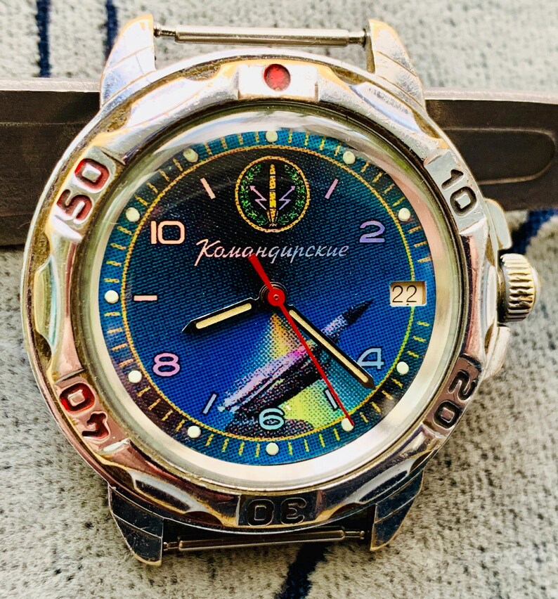 Op de afbeelding: Een vintage polshorloge met een zilverkleurige kast en een blauwe wijzerplaat. De wijzerplaat toont het Cyrillische woord "Командирские" en een raketafbeelding. Het horloge heeft een rode secondewijzer en lichtgevende uurmarkeringen.
