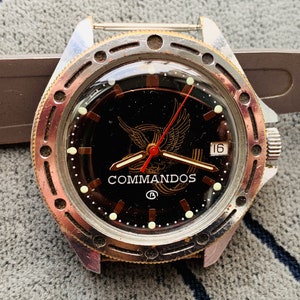 May include: A black and gold wristwatch with a silver strap. The watch face has a black background with a gold eagle logo and the word "COMMANDOS" printed below it. The watch has a date window at the 3 o'clock position and a red second hand.