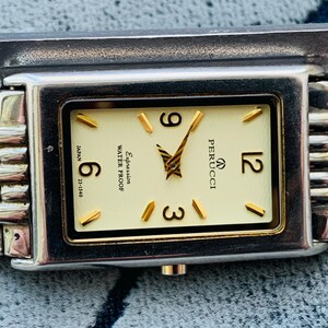 May include: A rectangular Perucci wristwatch with a cream-colored face, gold-tone hands and markers, and a silver and gold-tone band. The watch face displays the numbers 6, 9, and 12. The watch is water-resistant.