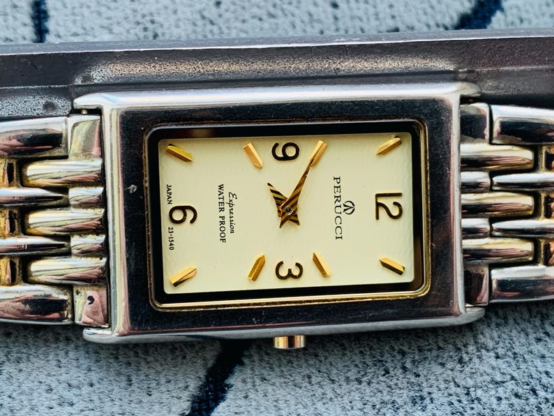 May include: A rectangular Perucci wristwatch with a cream-colored face, gold-tone hands and markers, and a silver and gold-tone band. The watch face displays the numbers 6, 9, and 12. The watch is water-resistant.