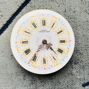 K&ouml;nnte beinhalten: Ein Vintage-Zifferblatt mit dem Text "HARDY A PETIT MARE". Das wei&szlig;e Zifferblatt hat schwarze r&ouml;mische Ziffern, goldene florale Akzente und filigrane Zeiger. Das Zifferblatt ist rund und scheint aus Porzellan oder einem &auml;hnlichen Material zu bestehen.