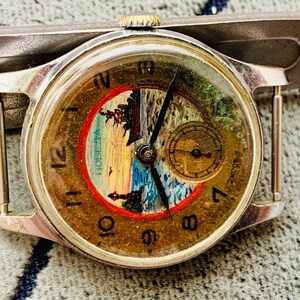 Puede incluir: Reloj de pulsera plateado vintage con esfera redonda. La esfera presenta un paisaje con un barco, un faro y la palabra "Победа" en un c&iacute;rculo rojo. El reloj tiene agujas negras y una peque&ntilde;a subesfera. El reloj est&aacute; sujeto a una correa plateada.