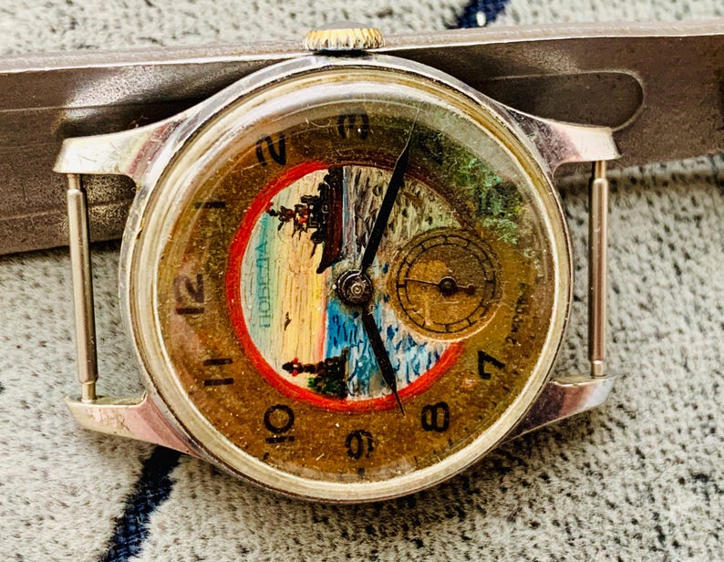 Puede incluir: Reloj de pulsera plateado vintage con esfera redonda. La esfera presenta un paisaje con un barco, un faro y la palabra "Победа" en un c&iacute;rculo rojo. El reloj tiene agujas negras y una peque&ntilde;a subesfera. El reloj est&aacute; sujeto a una correa plateada.