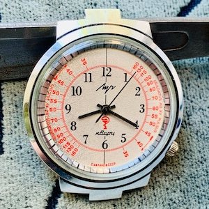 May include: A vintage silver-toned wristwatch with a white face and black hands. The watch features red numerals and markings, including the text "кварц" and "Сделано в СССР". The watch is set against a textured background.