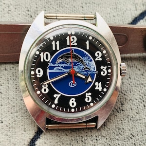 Puede incluir: Un reloj de pulsera vintage con caja plateada y esfera negra. La esfera presenta números blancos, un centro azul con un gráfico de delfín y el texto "Командирские". El reloj tiene un segundero rojo y una correa plateada.