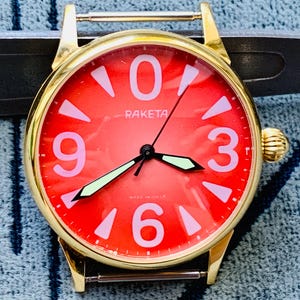 Puede incluir: Un reloj de pulsera Raketa en tono dorado con una esfera roja vibrante y grandes números de color rosa claro en las posiciones 0, 3, 6 y 9. El reloj tiene manecillas luminosas, una caja redonda y una corona lateral con el texto MADE IN USSR.