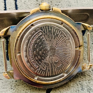 Op de afbeelding: Close-up van de achterkant van een vintage polshorloge. Het horloge heeft een goudkleurige bezel en een zilverkleurige achterkant met ge&euml;tste details. De achterkant van het horloge heeft Cyrillische tekst en een sunburst-ontwerp. Het horloge is bevestigd aan een metalen band.