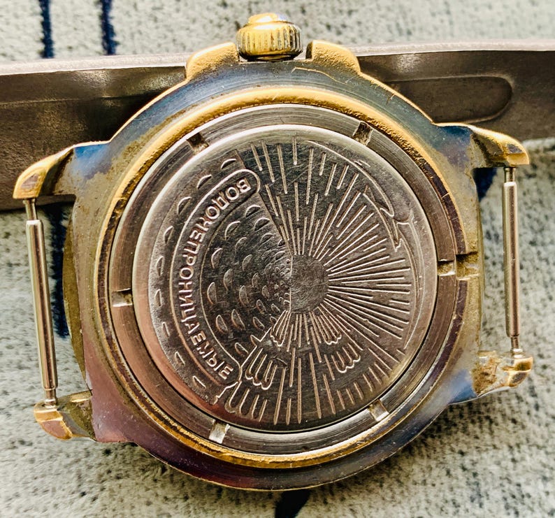 Op de afbeelding: Close-up van de achterkant van een vintage polshorloge. Het horloge heeft een goudkleurige bezel en een zilverkleurige achterkant met ge&euml;tste details. De achterkant van het horloge heeft Cyrillische tekst en een sunburst-ontwerp. Het horloge is bevestigd aan een metalen band.
