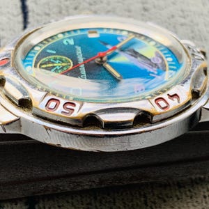 Op de afbeelding: Een vintage polshorloge met een zilverkleurige kast en een blauw en groene wijzerplaat. De wijzerplaat heeft een kompasontwerp en een rode secondewijzer. De bezel heeft goudkleurige markeringen. Een klassiek horloge.