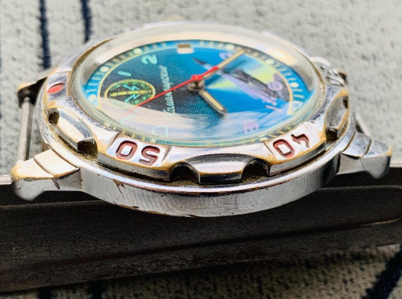 Op de afbeelding: Een vintage polshorloge met een zilverkleurige kast en een blauw en groene wijzerplaat. De wijzerplaat heeft een kompasontwerp en een rode secondewijzer. De bezel heeft goudkleurige markeringen. Een klassiek horloge.