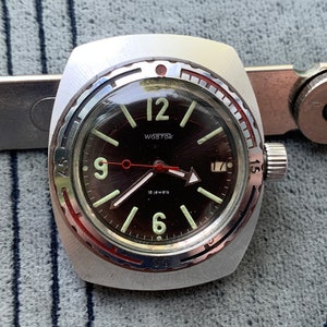 Puede incluir: Un reloj de pulsera plateado con una esfera negra y detalles rojos. El reloj tiene un bisel giratorio con números y un triángulo blanco en la posición de las 12 en punto. La esfera del reloj tiene números y manecillas blancas, y el texto "vostok 18 jewels" está impreso en la esfera.