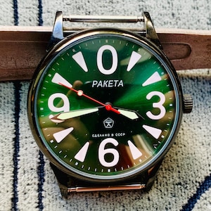 Peut inclure: Une montre-bracelet Raketa avec un cadran vert foncé soleillé, de grands chiffres blancs aux positions 0, 3, 6 et 9, et des index triangulaires blancs. Elle présente des aiguilles luminescentes, une trotteuse rouge et le texte "PAKETA" et "СДЕЛАНО В СССР".
