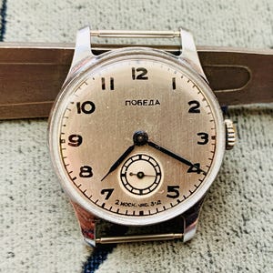 Peut inclure: Une montre-bracelet argentée vintage avec un cadran clair. La montre présente des aiguilles noires, des chiffres arabes et le mot "ПОБЕДА" en cyrillique. La montre est posée sur un fond métallique.