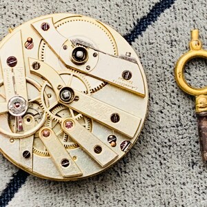 K&ouml;nnte beinhalten: Nahaufnahme eines Vintage-Uhrwerks mit filigranen Zahnr&auml;dern und Komponenten in Gold- und Silbert&ouml;nen. Ein goldfarbener Uhrenschl&uuml;ssel befindet sich rechts. Der Uhrmechanismus ist ein komplexes Beispiel f&uuml;r Handwerkskunst.
