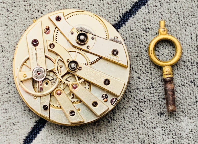 K&ouml;nnte beinhalten: Nahaufnahme eines Vintage-Uhrwerks mit filigranen Zahnr&auml;dern und Komponenten in Gold- und Silbert&ouml;nen. Ein goldfarbener Uhrenschl&uuml;ssel befindet sich rechts. Der Uhrmechanismus ist ein komplexes Beispiel f&uuml;r Handwerkskunst.