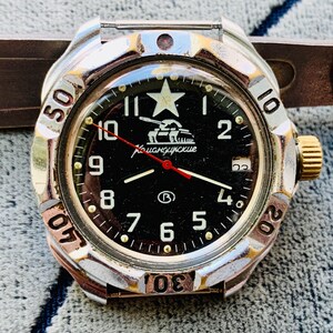May include: A vintage wristwatch with a black face, featuring white numerals and a star at the 12 o'clock position. The watch has a silver-tone bezel with minute markers and a gold-tone crown. The watch face includes the text "Командирские" and a tank graphic.