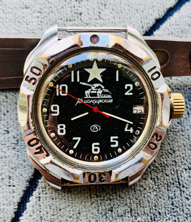 May include: A vintage wristwatch with a black face, featuring white numerals and a star at the 12 o'clock position. The watch has a silver-tone bezel with minute markers and a gold-tone crown. The watch face includes the text "Командирские" and a tank graphic.