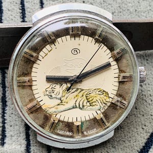 Puede incluir: Un reloj de pulsera vintage de plata con una esfera blanca y agujas negras. La esfera presenta una ilustración de tigre y el texto "СДЕЛАНО В СССР".