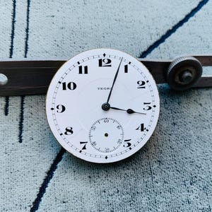 Puede incluir: Una esfera de reloj blanca con n&uacute;meros y manecillas negras, con un peque&ntilde;o dial de segundos. La palabra "TEGRA" es visible. Una herramienta de metal vintage est&aacute; colocada detr&aacute;s de la esfera.