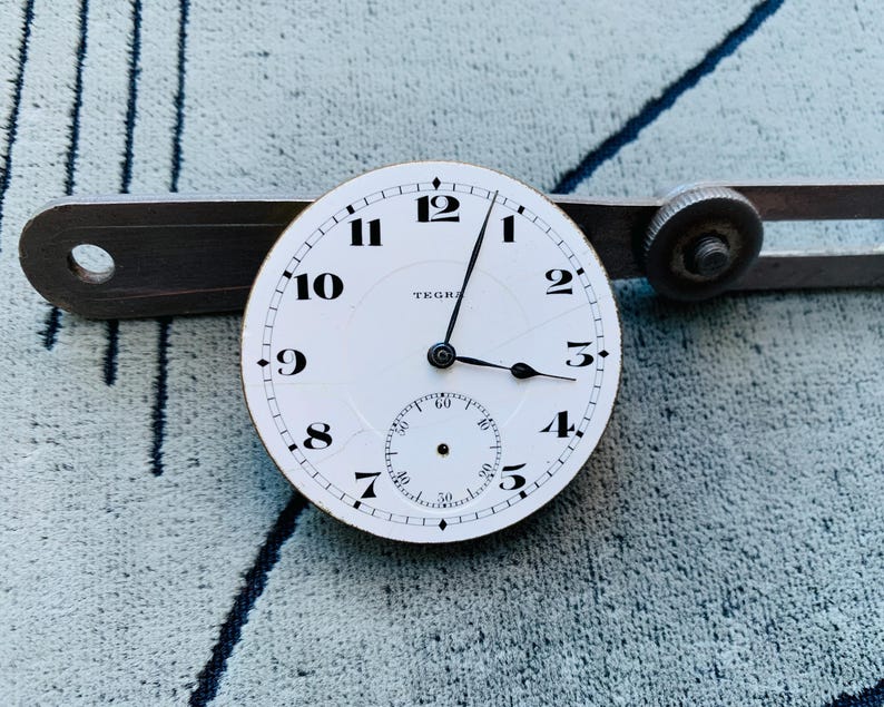 Puede incluir: Una esfera de reloj blanca con n&uacute;meros y manecillas negras, con un peque&ntilde;o dial de segundos. La palabra "TEGRA" es visible. Una herramienta de metal vintage est&aacute; colocada detr&aacute;s de la esfera.