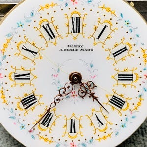 K&ouml;nnte beinhalten: Nahaufnahme eines Vintage-Zifferblatts mit r&ouml;mischen Ziffern und zarten floralen Akzenten. Das wei&szlig;e Zifferblatt hat schwarze Ziffern, goldfarbene Verzierungen und den Text "HARDY A PETIT MARS". Die Zeiger sind verziert und kupferfarben.