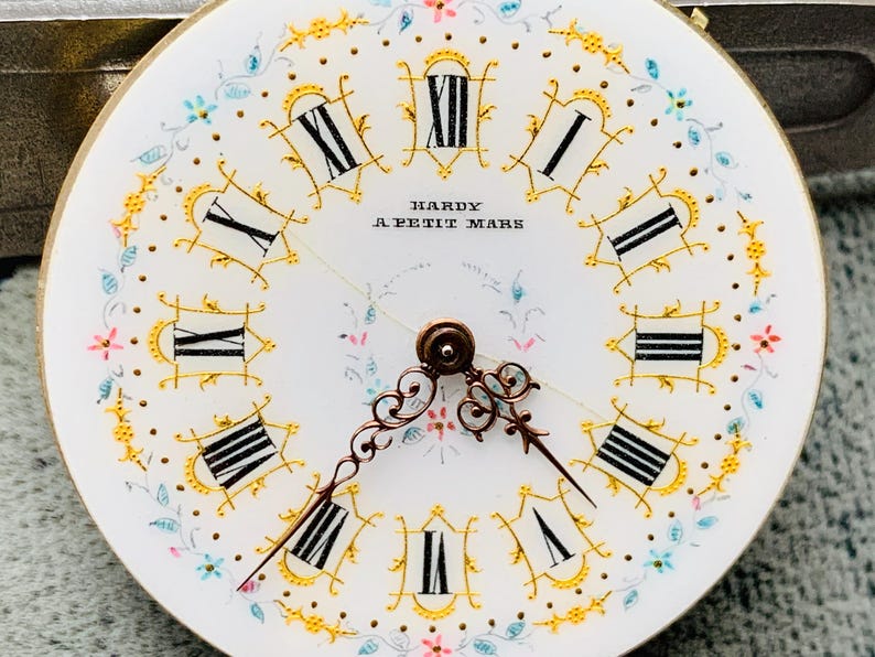 K&ouml;nnte beinhalten: Nahaufnahme eines Vintage-Zifferblatts mit r&ouml;mischen Ziffern und zarten floralen Akzenten. Das wei&szlig;e Zifferblatt hat schwarze Ziffern, goldfarbene Verzierungen und den Text "HARDY A PETIT MARS". Die Zeiger sind verziert und kupferfarben.