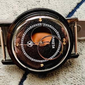 May include: Close-up of a black Raketa watch with a dark brown face. The watch features gold-colored accents and hands, with the text "PA KETA" and Cyrillic text. The watch is set on a light gray surface.