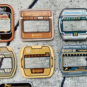 May include: A collection of vintage electronic watch faceplates in various colors, including red, brown, gold, and blue. Each plate features text in Cyrillic script and unique designs, with rectangular and oval shapes.