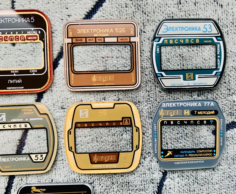 May include: A collection of vintage electronic watch faceplates in various colors, including red, brown, gold, and blue. Each plate features text in Cyrillic script and unique designs, with rectangular and oval shapes.