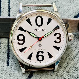 May include: A silver-toned Raketa wristwatch with a white face. The watch features large black numerals, black and green hands, and the word "RAKETA" in black. The second hand is red. The watch is a vintage style.