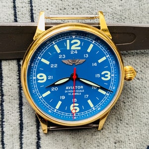 Puede incluir: Un reloj de aviador dorado con una esfera azul y marcas blancas. El reloj tiene una escala de 24 horas y dice "AVIATOR 24 HOUR SCALE 19 JEWELS".