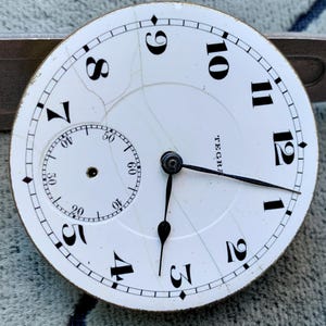Puede incluir: Primer plano de una esfera de reloj de bolsillo blanca con n&uacute;meros y manecillas negras. La esfera del reloj tiene una subesfera para los segundos y la palabra "TEGRA" est&aacute; impresa en la esfera. La esfera est&aacute; agrietada.