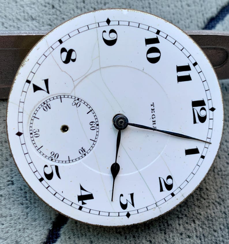 Puede incluir: Primer plano de una esfera de reloj de bolsillo blanca con n&uacute;meros y manecillas negras. La esfera del reloj tiene una subesfera para los segundos y la palabra "TEGRA" est&aacute; impresa en la esfera. La esfera est&aacute; agrietada.