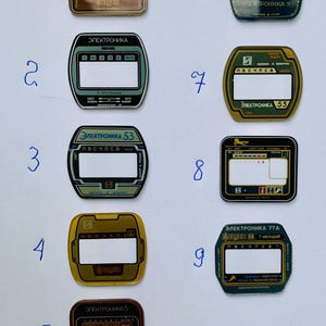 May include: A collection of nine vintage electronic device faceplates, each with a rectangular or oval display window. The faceplates feature text in Cyrillic script, with colors including gold, green, and black.