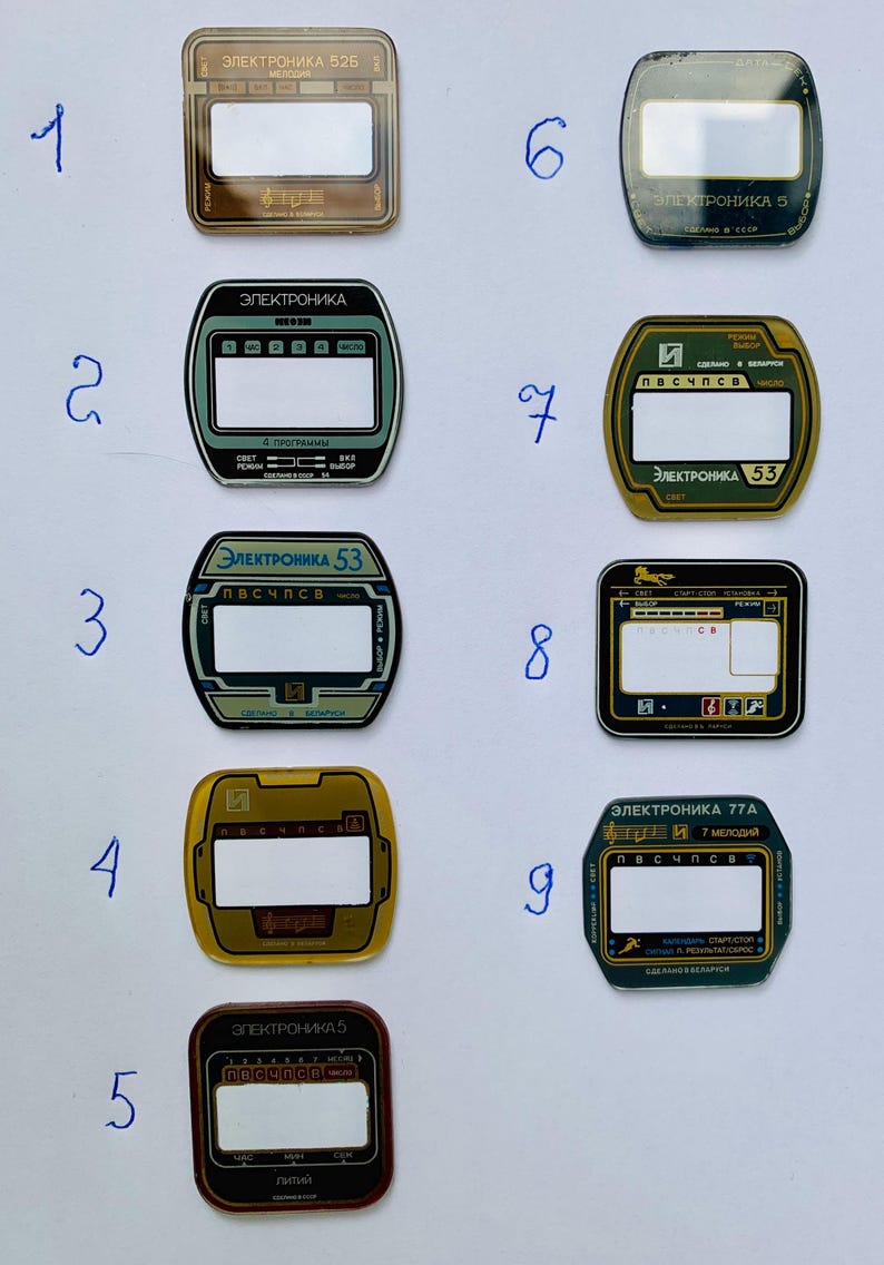 May include: A collection of nine vintage electronic device faceplates, each with a rectangular or oval display window. The faceplates feature text in Cyrillic script, with colors including gold, green, and black.