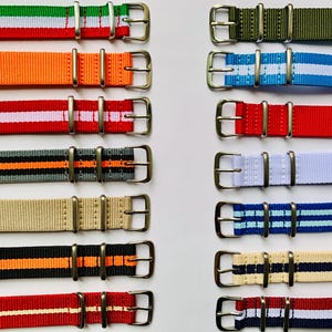 May include: A collection of twelve nylon watch straps arranged in two columns. Each strap features a silver-toned metal buckle and two matching metal keepers. The straps display various solid colors and striped patterns, including red, white, green, orange, blue, black, gray, and beige.
