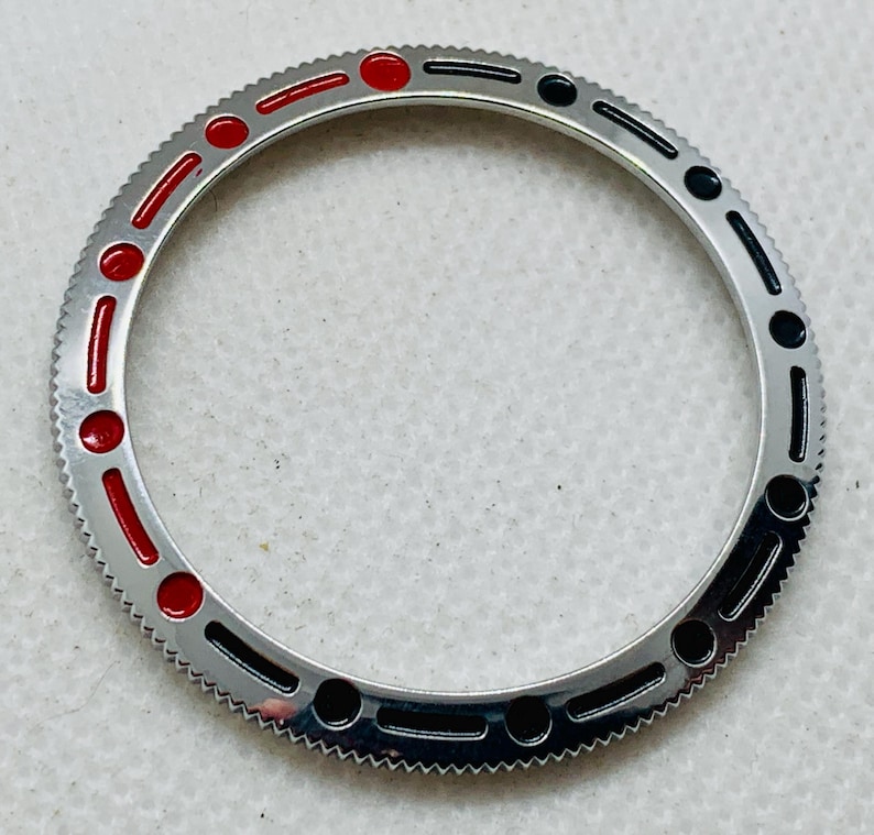 Op de afbeelding: Een zilveren horloge bezel met zwarte en rode markeringen. De bezel heeft een gekartelde rand en is ontworpen voor een horloge.