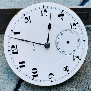Puede incluir: Primer plano de una esfera de reloj de bolsillo vintage. La esfera blanca presenta n&uacute;meros ar&aacute;bigos negros, un dial de segundos m&aacute;s peque&ntilde;o y la palabra "TEGRA". Las manecillas negras indican la hora. La esfera del reloj est&aacute; ligeramente envejecida.