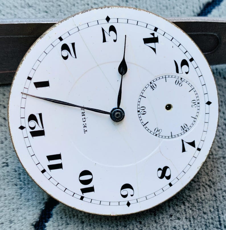 Puede incluir: Primer plano de una esfera de reloj de bolsillo vintage. La esfera blanca presenta n&uacute;meros ar&aacute;bigos negros, un dial de segundos m&aacute;s peque&ntilde;o y la palabra "TEGRA". Las manecillas negras indican la hora. La esfera del reloj est&aacute; ligeramente envejecida.