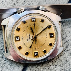 Puede incluir: Reloj de pulsera vintage Poljot de 18 rub&iacute;es con esfera dorada y caja plateada. El reloj presenta marcadores de hora y agujas rectangulares negros, con los n&uacute;meros 3, 6, 9 y 12 visibles. El reloj est&aacute; sobre un fondo texturizado.