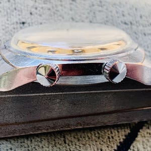 Puede incluir: Primer plano de un reloj de pulsera vintage con caja plateada y dos botones de corona texturizados. El reloj tiene un cristal transparente y abovedado y descansa sobre una superficie oscura y texturizada.