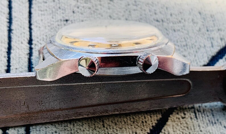 Puede incluir: Primer plano de un reloj de pulsera vintage con caja plateada y dos botones de corona texturizados. El reloj tiene un cristal transparente y abovedado y descansa sobre una superficie oscura y texturizada.