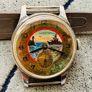 Puede incluir: Reloj de pulsera vintage con caja plateada y esfera redonda. La esfera presenta una escena n&aacute;utica con un faro, un barco y la palabra "Победа". El reloj tiene agujas negras y una peque&ntilde;a subesfera.