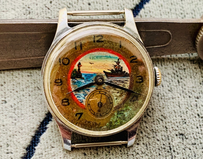 Puede incluir: Reloj de pulsera vintage con caja plateada y esfera redonda. La esfera presenta una escena n&aacute;utica con un faro, un barco y la palabra "Победа". El reloj tiene agujas negras y una peque&ntilde;a subesfera.