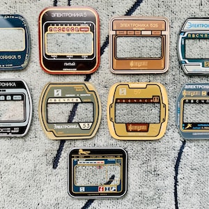 May include: A collection of vintage electronic watch faceplates in various colors, including black, gold, and blue. Each plate features a rectangular display window and Cyrillic text, with designs that include musical notes and numbers.